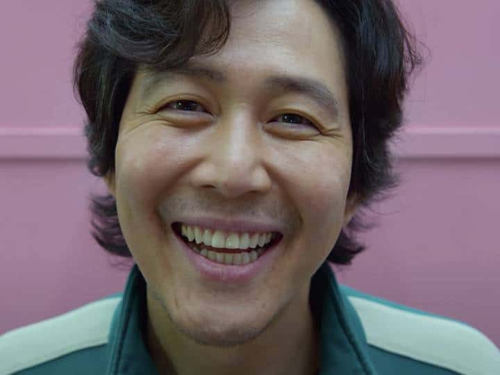 le visage d'un homme coréen souriant devant un mur rose, Squid games