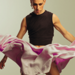 Sur fond gris, un homme avec un haut noir et une jupe rose