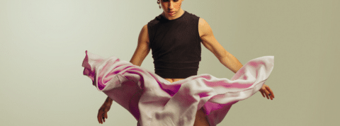 Comment les hommes jouent de plus en plus avec les codes de la mode Sur fond gris, un homme avec un haut noir et une jupe rose