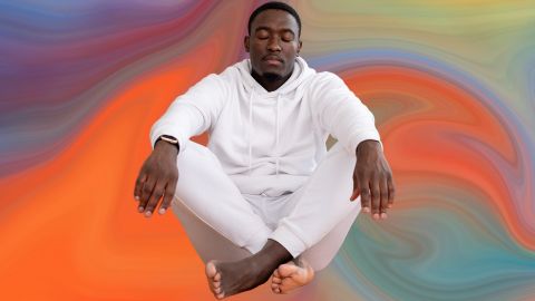 Un homme noir habillé en blanc assis en tailleur sur fond orange