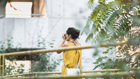 une femme qui prend une photo devant un ecalier d'hotel en béton brutaliste avec des plantes autours d'elle.