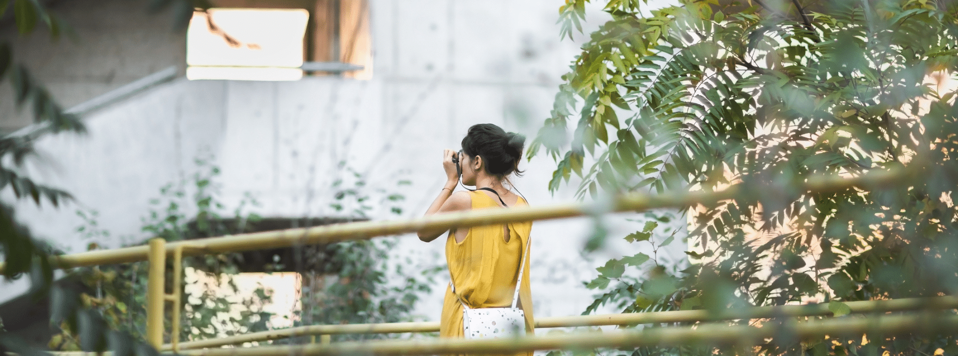 une femme qui prend une photo devant un ecalier d'hotel en béton brutaliste avec des plantes autours d'elle.