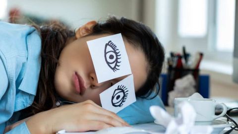 Femme dormant sur son bureau de travail, avec des post-it imitant les yeux ouverts
