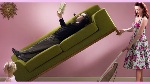 Un homme allongé sur un canapé et sa femme qui passe l'aspirateur