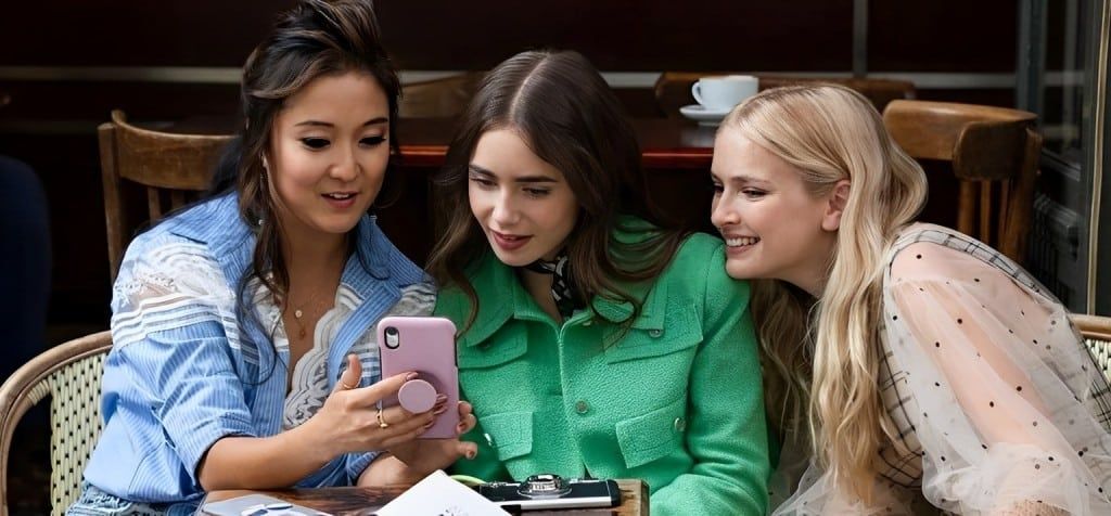 Emily in Paris et ses deux amies regardent un téléphone à la terrasse d'un café