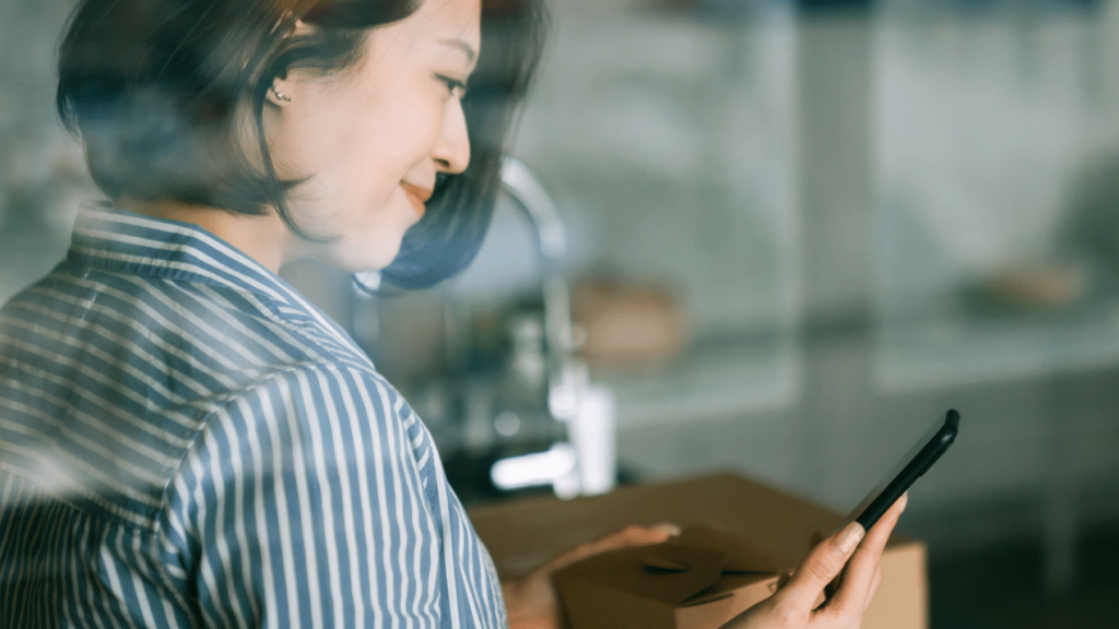 Femme asiatique souriante se tenant derrière une fenêtre, tenant son téléphone et gérant un système de notation depuis sa cuisine.