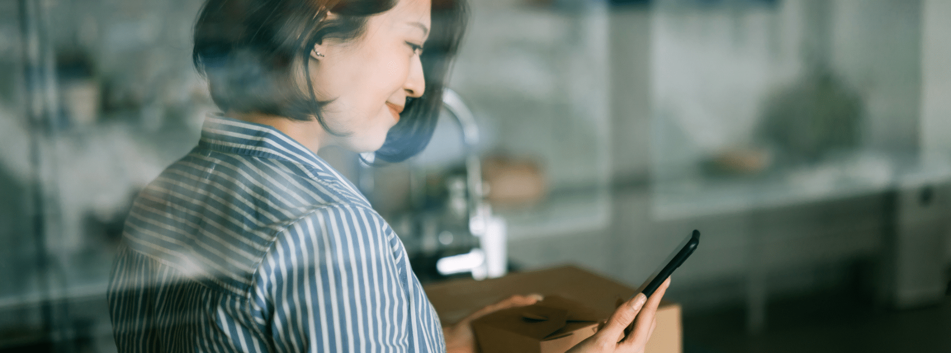 AsiaVision - L'ADN Femme asiatique souriante se tenant derrière une fenêtre, tenant son téléphone et gérant un système de notation depuis sa cuisine.