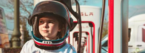 Read the fucking manual Un jeune enfant avec un casque et une tenue spatiale devant des bornes électriques Tesla