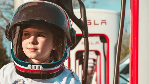 Un jeune enfant avec un casque et une tenue spatiale devant des bornes électriques Tesla