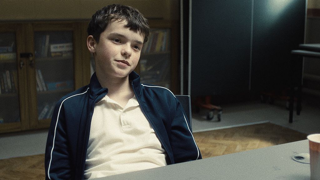 une jeune garçon assi. à une table l'air sur de lui.