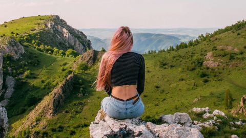 Une femme de dos aux longs cheveux roses devant un paysage montagneux