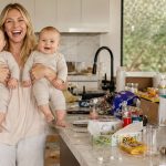 Une jeune maman souriante avec ses jumeaux dans les bras riant, dans une cuisine en bazar