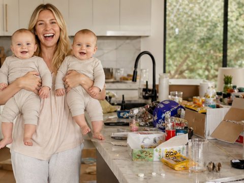 Une jeune maman souriante avec ses jumeaux dans les bras riant, dans une cuisine en bazar