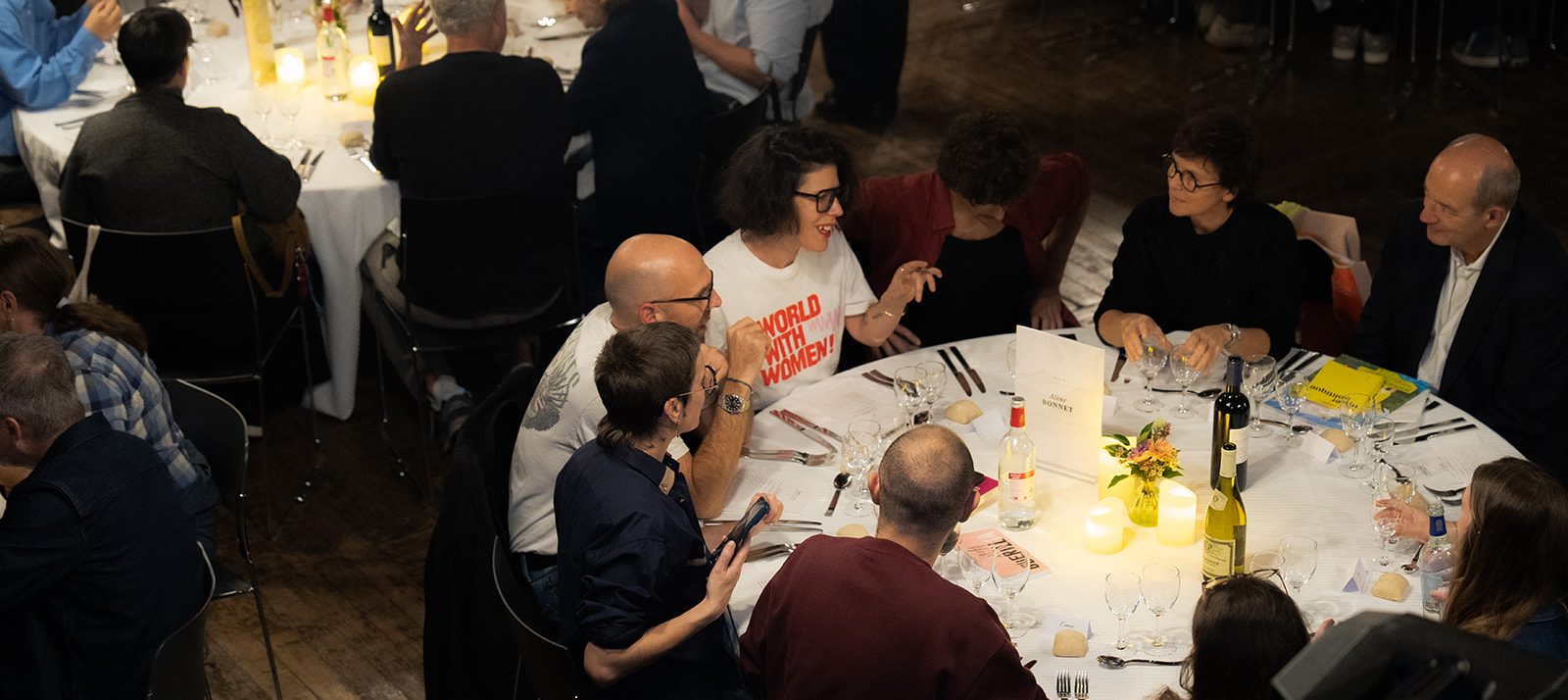 Dîner LADN Le Shift A round-table dinner scene with guests chatting, wine bottles, candles, and floral centerpieces at a formal event.