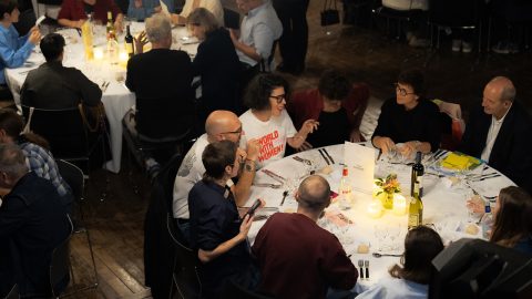 A round-table dinner scene with guests chatting, wine bottles, candles, and floral centerpieces at a formal event.