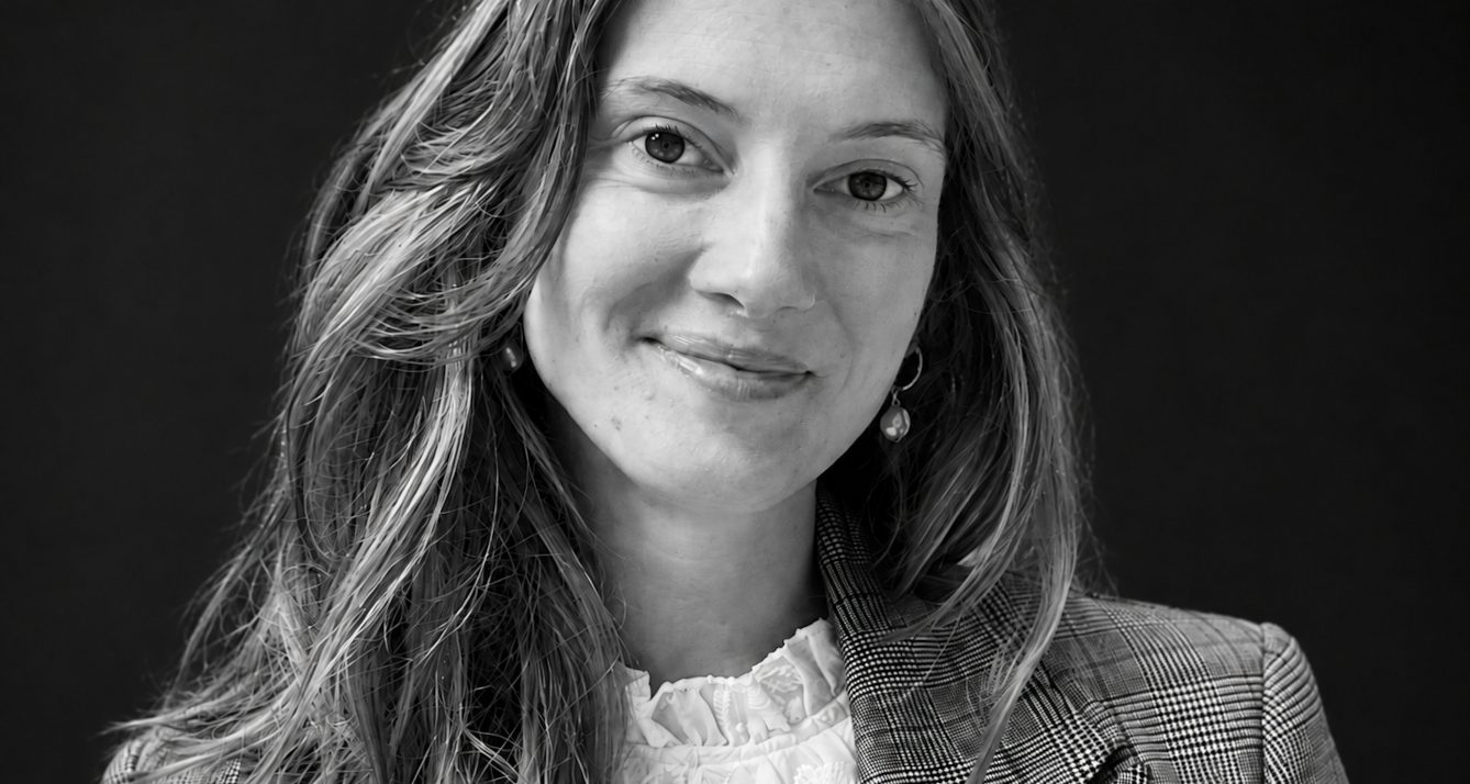 adn-l - L'ADN Black-and-white portrait of a smiling woman with wavy hair, wearing a plaid blazer and white blouse against a dark background.