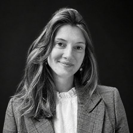 Black-and-white portrait of a smiling woman with wavy hair, wearing a plaid blazer and white blouse against a dark background.