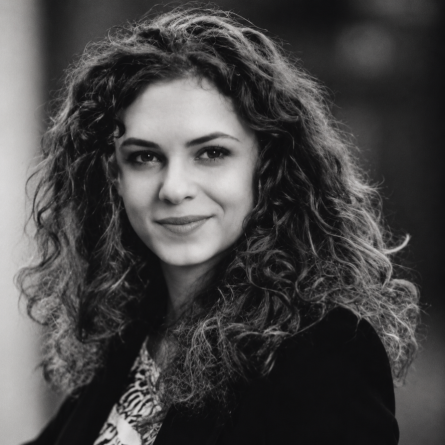 Head-and-shoulders black-and-white portrait of a woman with long curly hair, looking at the camera with a soft smile, wearing a dark jacket.