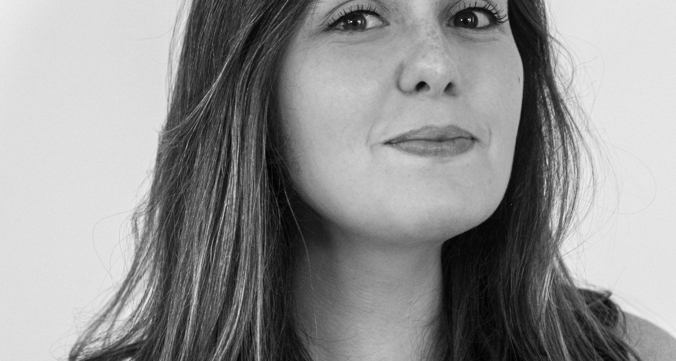 Close-up black-and-white portrait of a woman with shoulder-length hair, a subtle smile, wearing a white top and a small pendant necklace.