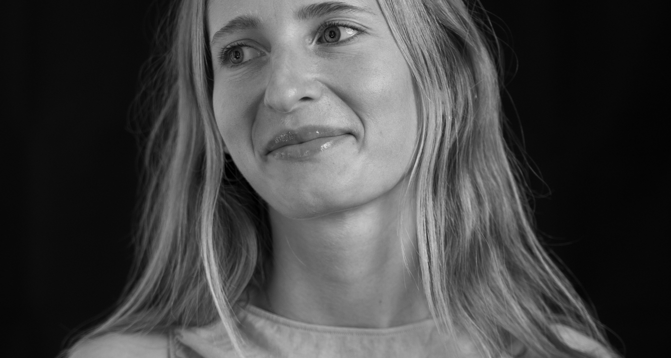 adn-r - L'ADN Black-and-white portrait of a smiling woman with long hair, looking slightly to the left, from the shoulders up against a dark background.