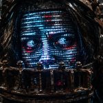 Cyborg head with glowing blue facial data pattern, held in rusty metal restraints in a dark dungeon.