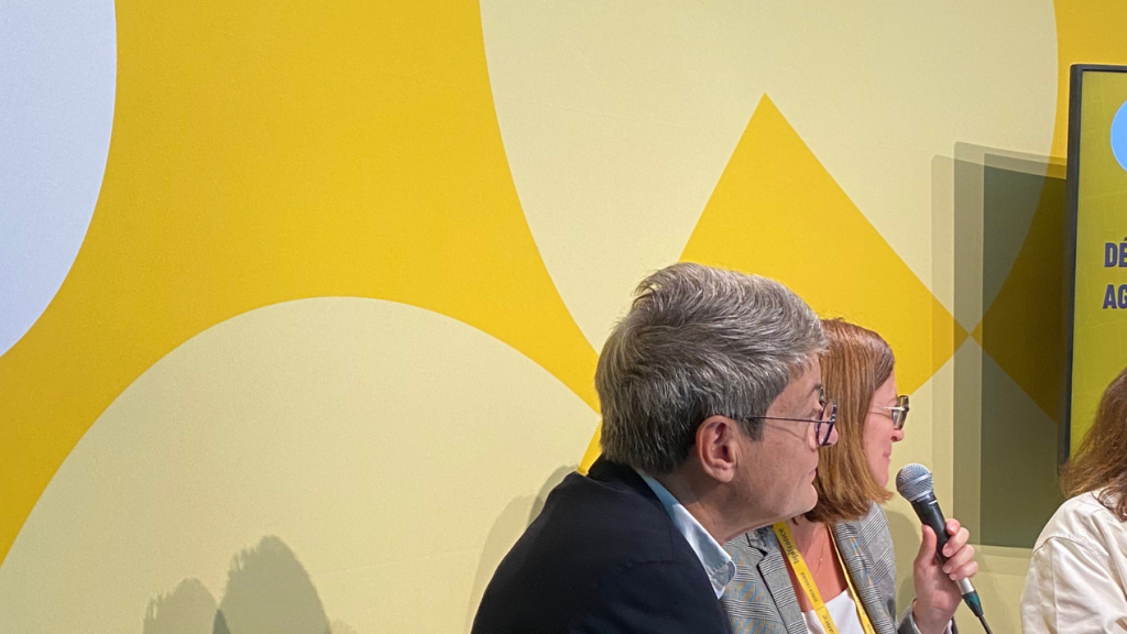 Three panelists sit with microphones on a bright yellow stage with ‘FRENCH CARE’ branding and a screen displaying session info in French.