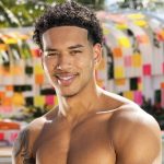 Smiling man with short curly hair, shirtless, in front of a colorful post-it-style art installation.
