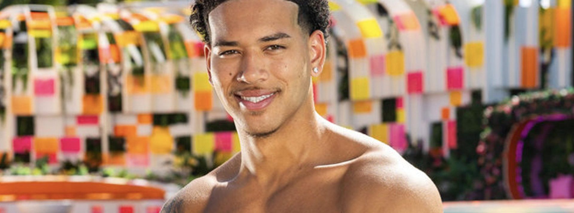 Smiling man with short curly hair, shirtless, in front of a colorful post-it-style art installation.
