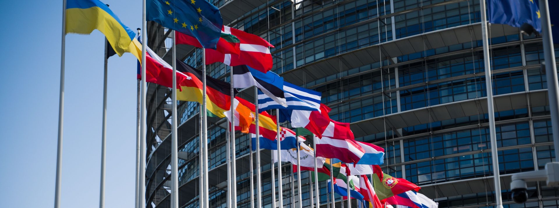 Row of colorful national flags on flagpoles in front of a modern glass office building against a blue sky.
